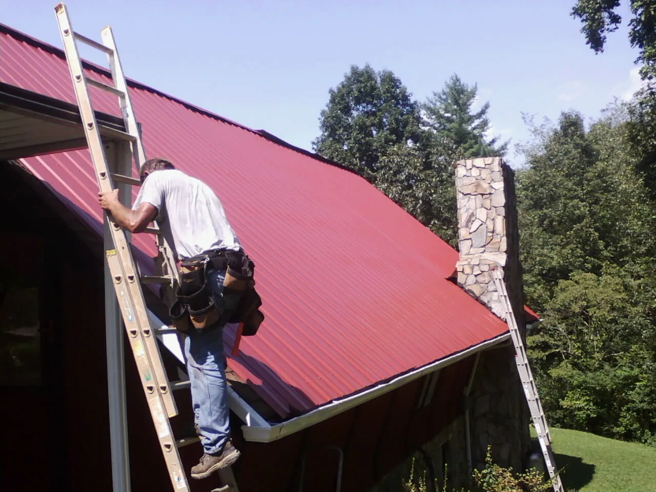 Roofer on a ladder beside a red metal roof and stone chimney — Soffit & Fascia Repair by Roofing Brothers in Mohrsville